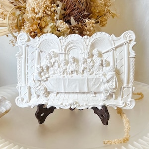 May include: White plaster relief sculpture depicting the Last Supper. The scene shows Jesus and his disciples gathered around a table, with a white tablecloth and a simple setting. The sculpture is framed by a decorative border and is mounted on a wooden stand.
