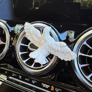 May include: A white dove car air vent decoration with outstretched wings. The dove is mounted on a black car dashboard with silver air vents. The dashboard has climate control buttons and text that reads "PASSENGER AIR BAG OFF".