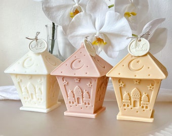 May include: Three decorative candle lanterns in the shape of a mosque with a crescent moon and stars. The lanterns are white, pink, and beige. The lanterns are made of wax and have a label that says "LMJ Candles".
