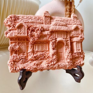 May include: A pink ceramic cottage figurine with a thatched roof, detailed brickwork, and a small garden. The figurine is on a wooden stand.