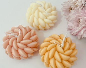May include: Three round candles with a unique leaf-like design. The candles are in shades of cream, peach, and yellow.