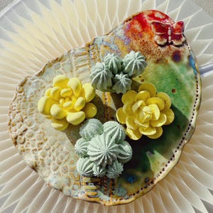 May include: A ceramic plate with a distinctive shape, adorned with yellow and green succulent sculptures. The plate has a textured surface, a butterfly detail, and a mix of colours, including red, green, and brown.
