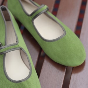 K&ouml;nnte beinhalten: Ein Paar gr&uuml;ne Mary Jane-Flats mit Riemen und grauem Besatz. Die Schuhe sind aus weichem Material und haben eine runde Spitze. Die Schuhe stehen auf einer braunen Holzoberfl&auml;che.