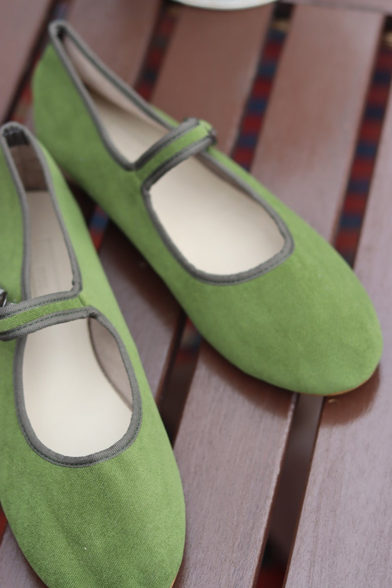 K&ouml;nnte beinhalten: Ein Paar gr&uuml;ne Mary Jane-Flats mit Riemen und grauem Besatz. Die Schuhe sind aus weichem Material und haben eine runde Spitze. Die Schuhe stehen auf einer braunen Holzoberfl&auml;che.