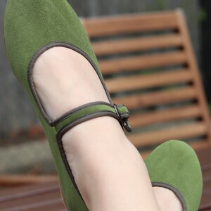 K&ouml;nnte beinhalten: Ein Paar gr&uuml;ne Mary Jane Ballerinas mit Riemen und Schnalle. Die Schuhe haben eine runde Spitze und eine dunkelgraue Einfassung. Die Schuhe sind aus einem weichen, Wildleder-&auml;hnlichen Material.