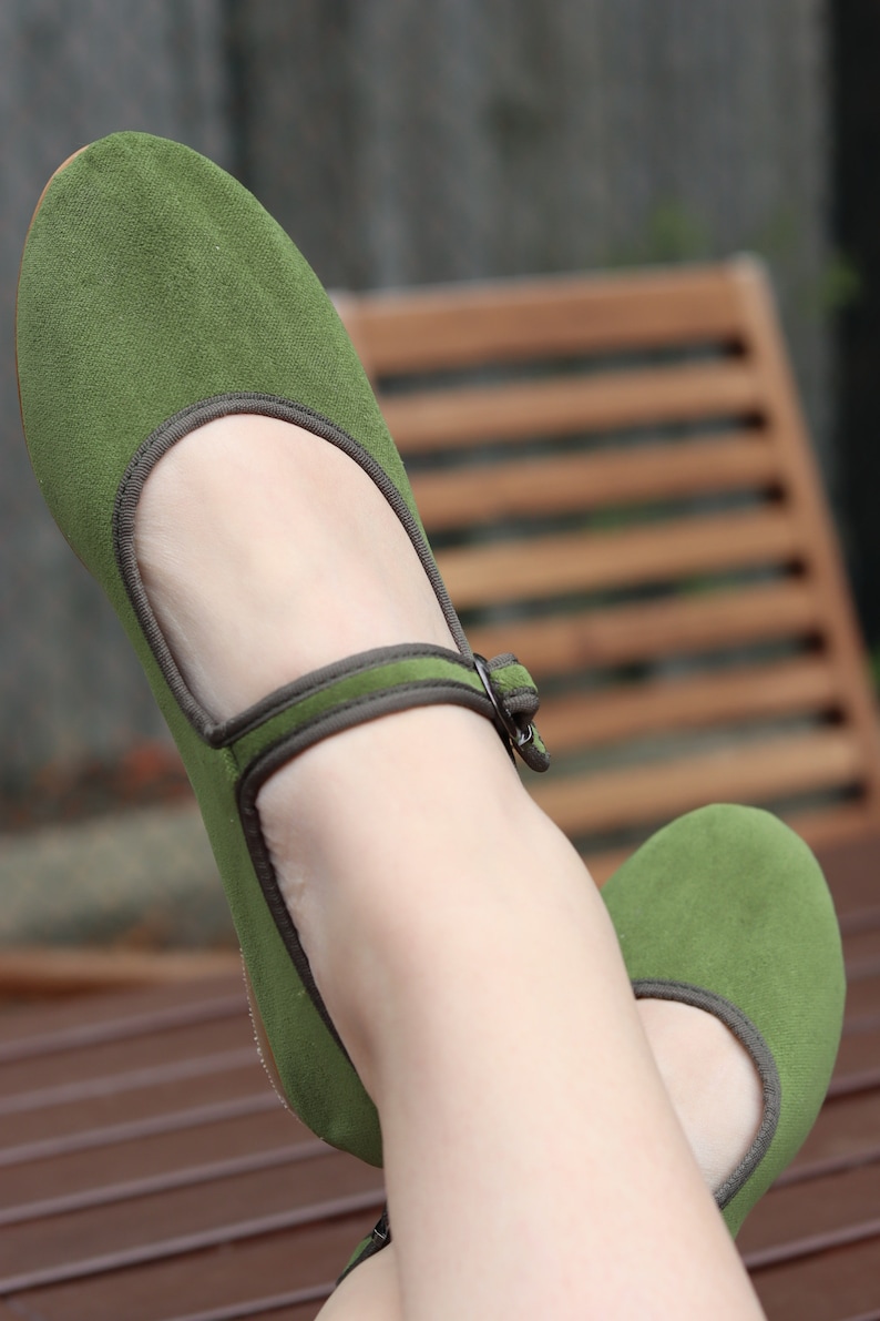 K&ouml;nnte beinhalten: Ein Paar gr&uuml;ne Mary Jane Ballerinas mit Riemen und Schnalle. Die Schuhe haben eine runde Spitze und eine dunkelgraue Einfassung. Die Schuhe sind aus einem weichen, Wildleder-&auml;hnlichen Material.