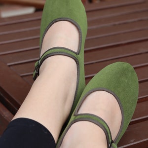 K&ouml;nnte beinhalten: Ein Paar gr&uuml;ne Mary Jane Schuhe mit Riemen und Schnalle. Die Schuhe haben eine runde Spitze und braune Zierleisten. Die Schuhe sind aus einem weichen, Wildleder-&auml;hnlichen Material.