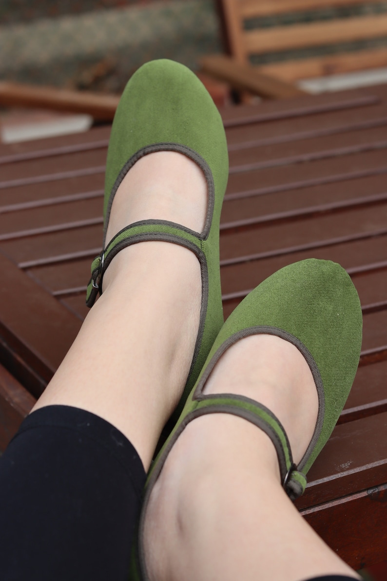 K&ouml;nnte beinhalten: Ein Paar gr&uuml;ne Mary Jane Schuhe mit Riemen und Schnalle. Die Schuhe haben eine runde Spitze und braune Zierleisten. Die Schuhe sind aus einem weichen, Wildleder-&auml;hnlichen Material.