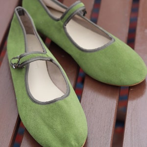 K&ouml;nnte beinhalten: Ein Paar gr&uuml;ne Mary Jane-Flats mit Riemen und Schnallenverschluss. Die Schuhe haben eine runde Spitze und eine kontrastierende graue Einfassung. Die Schuhe sind aus einem weichen, strukturierten Material gefertigt und liegen auf einer Holzoberfl&auml;che.