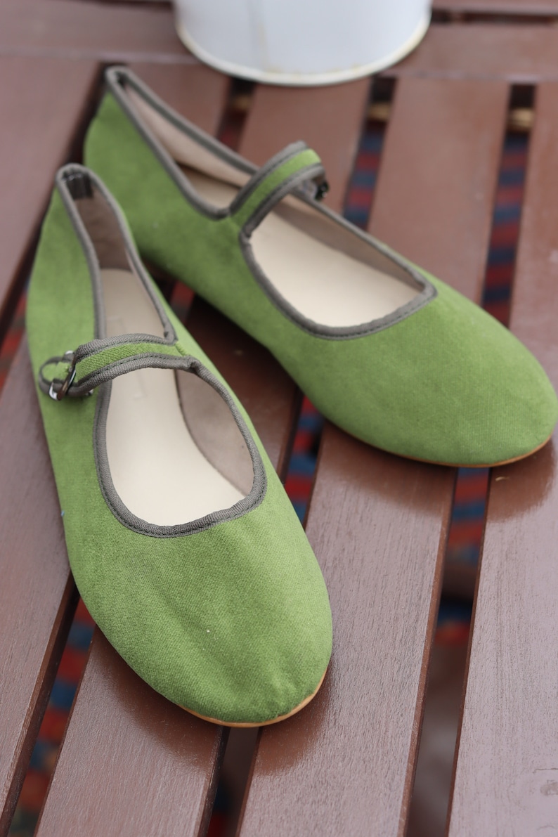 K&ouml;nnte beinhalten: Ein Paar gr&uuml;ne Mary Jane-Flats mit Riemen und Schnallenverschluss. Die Schuhe haben eine runde Spitze und eine kontrastierende graue Einfassung. Die Schuhe sind aus einem weichen, strukturierten Material gefertigt und liegen auf einer Holzoberfl&auml;che.