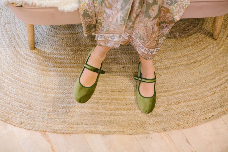 K&ouml;nnte beinhalten: Olivgr&uuml;ne Mary Jane-Ballerinas mit Riemen und Schnallenverschluss. Die Schuhe sind aus einem weichen, samtigen Material gefertigt. Die Schuhe werden mit einem gemusterten Kleid getragen und liegen auf einem gewebten Jute-Teppich.