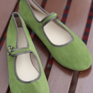 K&ouml;nnte beinhalten: Ein Paar gr&uuml;ne Mary Jane-Schuhe aus Wildleder mit Riemen und Schnalle. Die Schuhe haben eine runde Spitze und eine graue Einfassung. Sie liegen auf einer braunen Holzoberfl&auml;che.