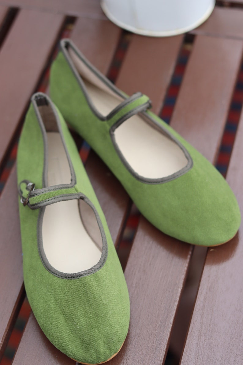 K&ouml;nnte beinhalten: Ein Paar gr&uuml;ne Mary Jane-Schuhe aus Wildleder mit Riemen und Schnalle. Die Schuhe haben eine runde Spitze und eine graue Einfassung. Sie liegen auf einer braunen Holzoberfl&auml;che.