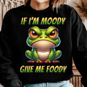 May include: Black sweatshirt featuring a green cartoon frog with angry eyes and the text "If I'm Moody Give Me Foody".