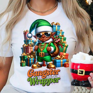 May include: White t-shirt featuring a cartoon illustration of a green elf wearing a Santa hat and holding a bag of presents. The elf is surrounded by wrapped presents and the text "Gangster Wrapper" is printed below the image.