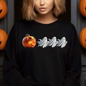 Puede incluir: Sudadera negra con un diseño de Halloween. El diseño incluye una calabaza de dibujos animados con una amplia sonrisa y tres figuras de fantasmas blancos. Calabazas naranjas talladas en el fondo.