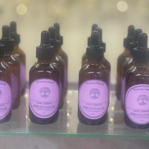 Puede incluir: Una colección de botellas de vidrio marrón con cuentagotas negros, cada una con una etiqueta rosa. La etiqueta dice "Glow Goddess Third Eye Facial Oil" con un diseño de árbol. Las botellas están en un estante de vidrio.