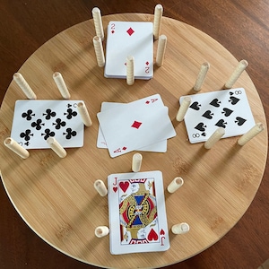 May include: A wooden game board with a circular shape and a light brown finish.  The board has eight slots around the edge, each with a wooden dowel.  There are five playing cards placed in the center of the board.  The cards are a Jack of Hearts, an Ace of Diamonds, an Eight of Spades, a Two of Diamonds, and a Queen of Clubs.