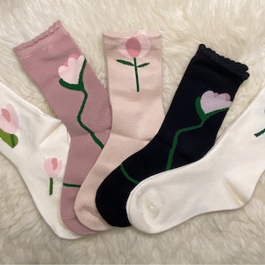 Könnte beinhalten: Fünf Paar Socken mit Blumenmustern. Die Socken sind in verschiedenen Farben, darunter Weiß, Rosa, Schwarz und Hellrosa. Jede Socke zeigt eine einzelne Blume mit einem grünen Stiel und Blättern.