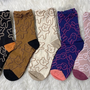 Könnte beinhalten: Fünf Paar Socken mit verschiedenen Mustern. Zwei Paar zeigen braune Teddybären auf schwarzem und braunem Hintergrund. Drei Paar zeigen rosa Hasen auf weißem, blauem und rosa Hintergrund.