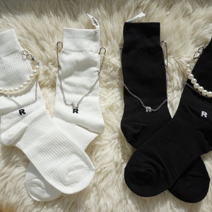 Peut inclure: Deux paires de chaussettes, une blanche et une noire, avec un détail de perles et de chaîne. Chaque chaussette a une épingle de sûreté et la lettre "R" brodée sur le côté.