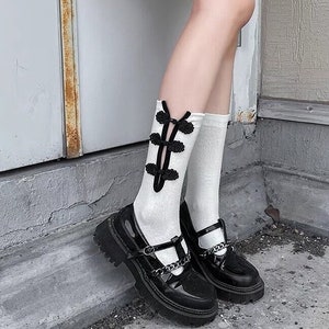 Könnte beinhalten: Schwarze Lack-Mary-Jane-Schuhe mit klobiger Sohle und silbernen Kettendetails. Die Schuhe werden mit weißen Socken mit schwarzen Zierakzenten kombiniert. Die Schuhe stehen vor einem Betonhintergrund.
