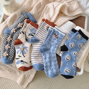 Könnte beinhalten: Ein Set mit sechs bunten Socken mit verschiedenen Mustern, darunter florale, geometrische und gestreifte Designs. Die Socken liegen auf einer weißen Strickdecke.