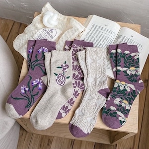 Könnte beinhalten: Fünf Paar Socken mit Blumenmustern in Violett- und Cremetönen. Die Socken liegen auf einer Holzoberfläche.