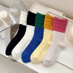 Op de afbeelding: Zes paar kleurrijke sokken met witte geribbelde boorden. De sokken zijn in verschillende kleuren, waaronder zwart, groen, geel, roze en blauw. De sokken zijn allemaal gevouwen en in een rij gerangschikt.