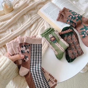 Könnte beinhalten: Eine Auswahl an gemusterten Socken in verschiedenen Farben, darunter Rosa, Braun, Grün und Schwarz. Die Socken weisen Blumen-, Hahnentritt- und Stickmuster auf. Einige Socken haben Rüschenkanten. Die Socken werden auf einer weißen Oberfläche mit einem Buch und einer Strickdecke im Hintergrund präsentiert.