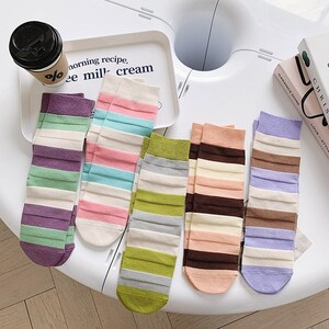 Könnte beinhalten: Sechs Paar gestreifte Socken in verschiedenen Farben, darunter Lila, Grün, Rosa, Blau, Braun und Grau. Die Socken liegen auf einer weißen Oberfläche.