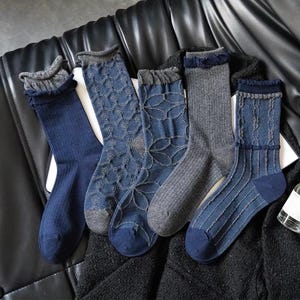 Könnte beinhalten: Eine Sammlung von fünf Paar Socken in verschiedenen Blau- und Grautönen. Die Socken weisen unterschiedliche Muster auf, darunter gerippte, florale und strukturierte Designs. Jedes Paar hat ein kontrastierendes Bündchen.
