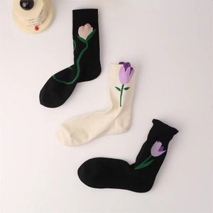 Puede incluir: Tres pares de calcetines con un diseño floral. Un par es negro con una flor morada, un par es blanco con una flor morada y un par es negro con una flor rosa.