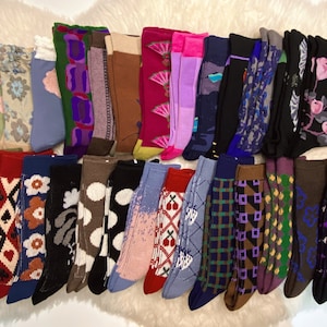Könnte beinhalten: Eine Sammlung von 28 bunten Socken mit verschiedenen Mustern, darunter geometrische, florale und abstrakte Designs. Die Socken sind in einer Reihe auf einer weißen, flauschigen Oberfläche angeordnet.