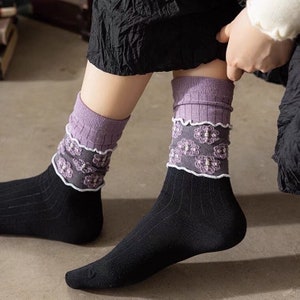 Könnte beinhalten: Ein Paar schwarze Socken mit violett-weißem Blumenbesatz. Die Socken sind gerippt und haben ein zartes Spitzendetail.