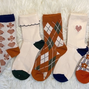 Könnte beinhalten: Fünf Paar bunte Socken mit verschiedenen Mustern. Die Socken sind weiß mit braunen, grünen und blauen Akzenten. Die Muster umfassen Spielkarten, Rauten, Herzen und Blumen.