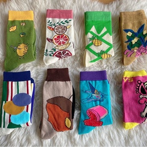 Könnte beinhalten: Acht Paar bunte Socken mit verschiedenen Mustern, darunter Zitronen, Ananas, ein Vogel und eine Comicfigur. Die Socken sind in zwei Reihen auf einer weißen, flauschigen Oberfläche angeordnet.