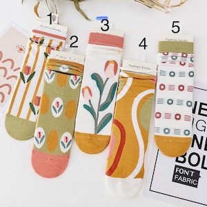 Könnte beinhalten: Eine Sammlung von fünf Paar Socken mit Mustern. Designs umfassen Streifen, Blumenmuster und geometrische Muster in Farben wie Grün, Rosa und Gelb. Jedes Paar ist mit "Fashion Socks" beschriftet. Eine Karte mit Regenbogen- und Augengrafiken ist ebenfalls vorhanden.