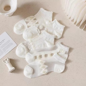 Op de afbeelding: Een verzameling van vijf witte enkelsokken met verschillende kant- en textuurpatronen. De sokken zijn gerangschikt op een lichtgekleurd oppervlak, met een kleine decoratieve zuil en een stuk papier op de achtergrond.