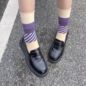 Könnte beinhalten: Ein Paar schwarze Lederschuhe mit dicker Sohle und einem Riemen über dem Oberteil. Die Schuhe werden mit Socken getragen, die ein pinkfarbenes, blaues und violettes Streifenmuster haben.