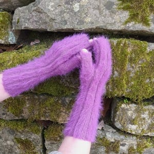 May include: A pair of vibrant purple fingerless gloves. The gloves are made of a soft, fuzzy material and feature a ribbed design on the back of the hand. The background is a stone wall covered in green moss.