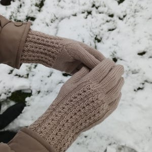 May include: A pair of beige knitted gloves with a textured pattern on the back of the hand and along the wrist. The gloves are made of a soft, warm material, perfect for cold weather. The background is a snowy scene.