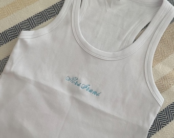 May include: A white ribbed tank top with "Mrs. Front" embroidered in light blue, alongside a white cowboy hat, fluffy slippers with pearl accents, and a white cosmetic bag with "BRIDE" spelled out in pearls. A decorative box with "BRIDE" is also visible.