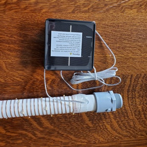 May include: A solar powered string of lights with a white plastic tube and a small solar panel. The panel has a warning label with instructions for use. The lights are coiled up and ready to be strung.