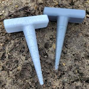 May include: Two gray T-shaped soil depth gauges with inch markings. One is speckled, the other solid. The gauges are on a bed of soil, with measurements from 1/2" to 4".