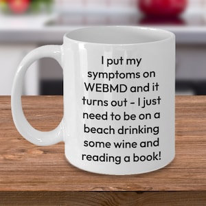 May include: White ceramic coffee mug with black text that reads "I put my symptoms on WEBMD and it turns out - I just need to be on a beach drinking some wine and reading a book!"