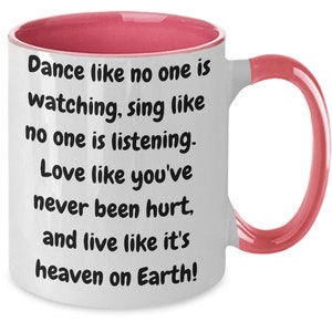 May include: White ceramic coffee mug with a pink rim and handle. The mug has a quote printed on it: "Dance like no one is watching, sing like no one is listening. Love like you've never been hurt, and live like it's heaven on Earth!"