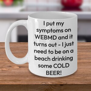 May include: White ceramic coffee mug with black text that reads "I put my symptoms on WEBMD and it turns out - I just need to be on a beach drinking some COLD BEER!"