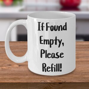 Puede incluir: Taza de cerámica blanca con asa curvada. La taza presenta el texto "If Found Empty, Please Refill!" en letras negras y en negrita. La taza está sobre una superficie de madera.