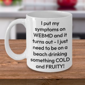 May include: White ceramic coffee mug with black text that reads "I put my symptoms on WEBMD and it turns out - I just need to be on a beach drinking something COLD and FRUITY!"